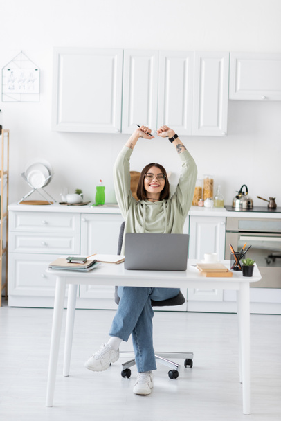 Excited freelancer in eyeglasses showing yes gesture near laptop and notebooks in kitchen  - Foto, Bild