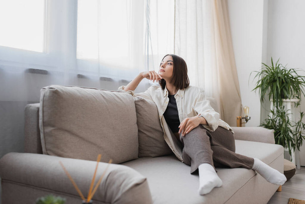 Junge Frau in lässiger Kleidung schaut weg, während sie zu Hause auf der Couch sitzt  - Foto, Bild