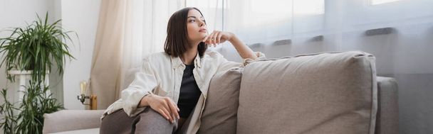 Brunette woman in casual clothes looking away while sitting on couch at home, banner  - Photo, image
