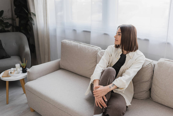 Mladá brunetka žena se usmívá, zatímco sedí na gauči v obývacím pokoji  - Fotografie, Obrázek
