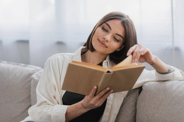 Pleased tattooed woman reading blurred book in living room  - Φωτογραφία, εικόνα