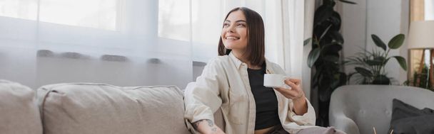 Overjoyed woman holding cup of coffee while sitting on couch at home, banner  - Foto, immagini
