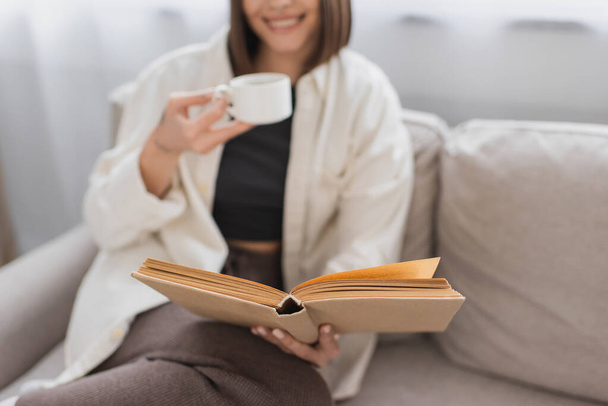 Cropped view of blurred woman holding coffee cup and reading book on couch at home  - Fotoğraf, Görsel