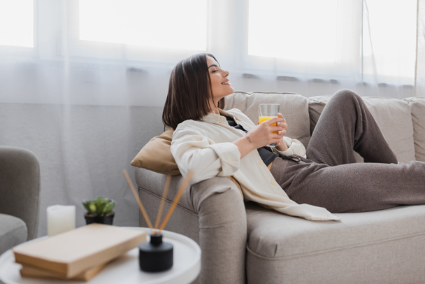 Vue latérale de la femme heureuse tenant un verre de jus d'orange sur le canapé à la maison  - Photo, image