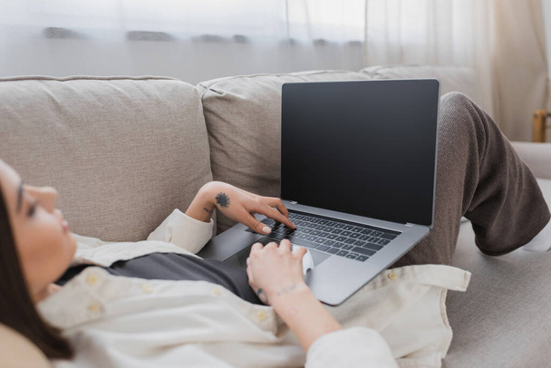 Freelancer tatuado borroso usando computadora portátil con pantalla en blanco en el sofá en casa  - Foto, imagen