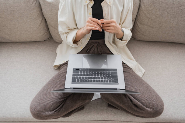 Cropped view of tattooed woman in casual clothes holding laptop while sitting on couch at home  - Fotoğraf, Görsel