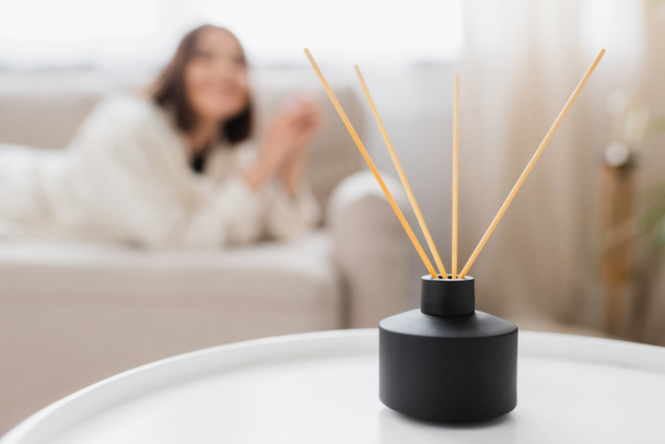 Bamboo sticks in aroma diffuser on coffee table near blurred woman in living room  - Photo, Image