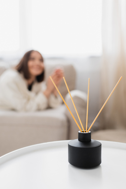 Aroma diffuser with bamboo sticks on coffee table near blurred woman in living room - Photo, Image
