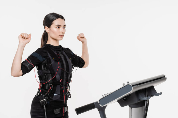 Γυμναστήριο κορίτσι στο EMS κοστούμι κάνει προπόνηση αθλητισμού κατάρτισης που χρησιμοποιεί ηλεκτρικά ερεθίσματα για την τόνωση των μυών σε λευκό φόντο. Αθλητική εκπαίδευση σε ηλεκτρική στολή μυϊκής διέγερσης - Φωτογραφία, εικόνα