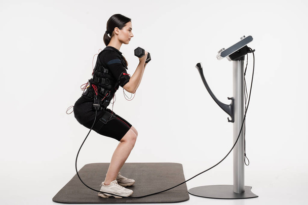 Fit girl does squats with dumbbells physical exercises in EMS suit that uses electrical impulses to stimulate muscles on white background. Sport training in electrical muscle stimulation suit - Foto, Bild