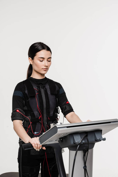 Girl trainer is setting up EMS device before training. Set up EMS device for workout sport training in electrical muscle stimulation suit on white background - Foto, Imagen