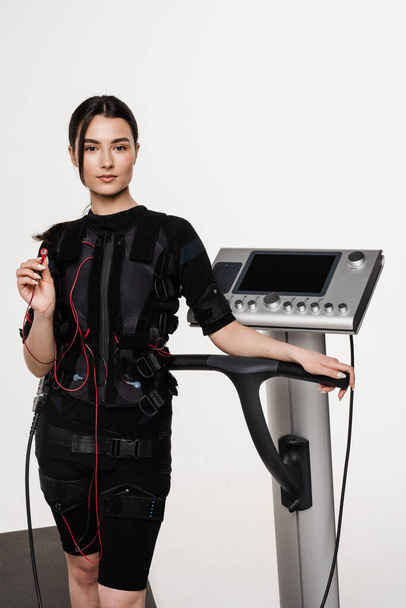 Fit dívka v EMS obleku s kontaktním prvkem v ruce, který používá elektrické impulsy stimulovat svaly na bílém pozadí. Sportovní trénink v elektrickém svalovém stimulačním obleku - Fotografie, Obrázek