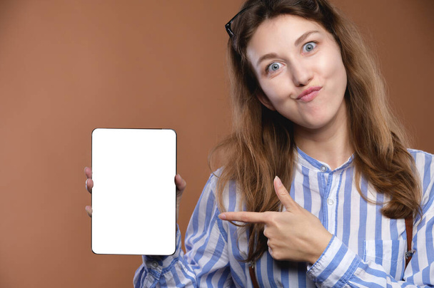 A grimace Caucasian young business style woman holds an election tablet with a cut-out screen in her hands. Funny business woman presenting app on tablet screen. - Foto, Imagem