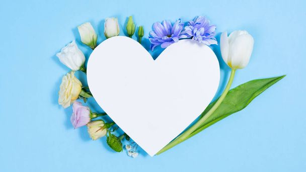 Φυσικά λευκά και μπλε-βιολετί λουλούδια κοντά στην καρδιά του λευκού χαρτιού. Ευχετήρια κάρτα για τα γενέθλια, την ημέρα της μητέρας, την ημέρα της γυναίκας ή άλλη περίσταση. Αντιγραφή χώρου. Επίπεδη τοποθέτηση. - Φωτογραφία, εικόνα