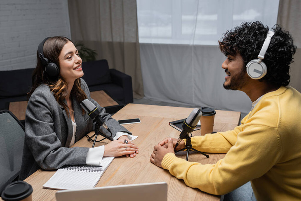 happy and charming radio host in headphones and young curly indian man in yellow jumper talking near microphones, paper cups, smartphone and notebooks in professional studio - Photo, Image