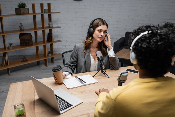 tattooed brunette interviewer in headphones holding takeaway drink and looking at blurred indian man near laptop, microphones, notebook and mobile phone on table in broadcasting studio - Foto, imagen