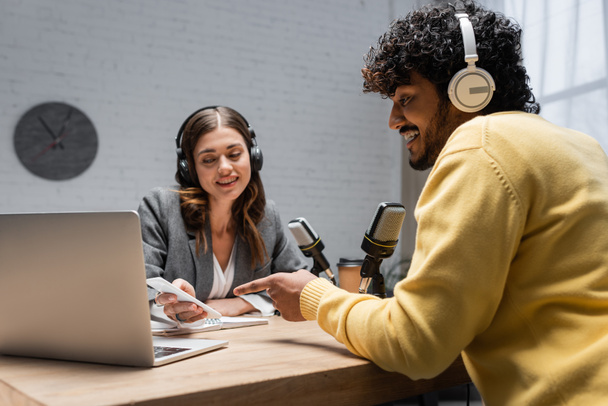 charming radio host in grey blazer showing smartphone to young indian man in headphones and yellow jumper pointing at laptop near professional microphones in broadcasting studio - Foto, Imagen
