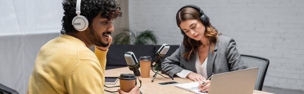 curly and happy indian man in headphones and yellow jumper sitting with paper cup near laptop and radio host writing in notebook close to smartphone with blank screen in broadcasting studio, banner - Photo, Image