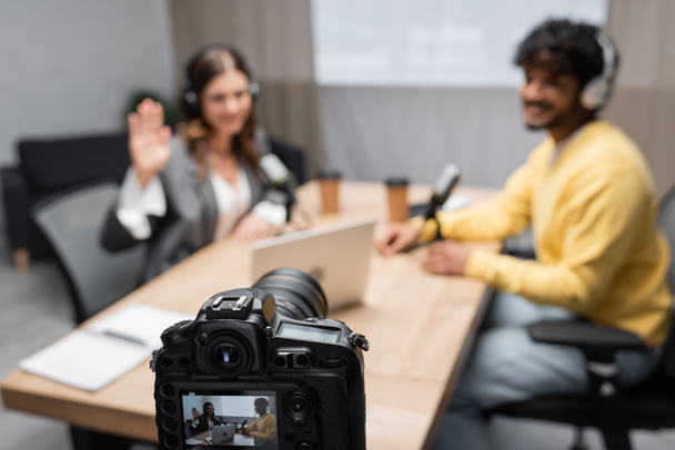 ammatillinen digitaalikamera tallennus haastattelu nainen heiluttaa käsi ja nuori intialainen vieras istuu pöydässä kannettava tietokone ja hämärtynyt paperi kupit lähetykset studio - Valokuva, kuva