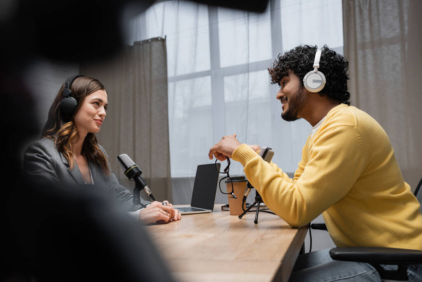 zijaanzicht van glimlachende en krullende indiaanse man in hoofdtelefoon en gele jumper praten met brunette interviewer in de buurt van laptop met leeg scherm en koffie te gaan in radio studio op wazig voorgrond - Foto, afbeelding