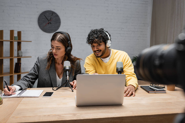 brunette radio host in headphones writing in notebook close to positive indian colleague looking at laptop near professional microphones and smartphone with blank screen in studio - 写真・画像