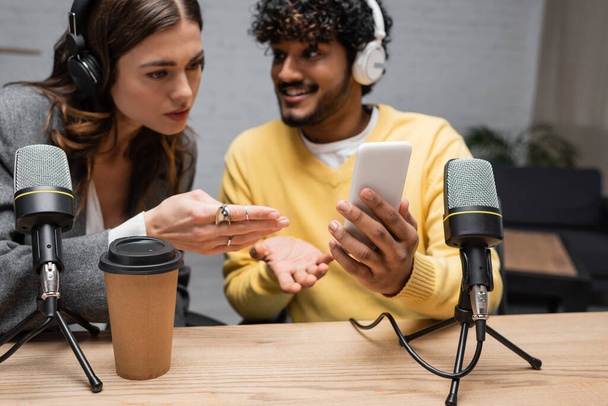 serious radio host in headphones pointing at mobile phone in hands of positive indian colleague in yellow jumper near coffee to go and microphones in broadcasting studio - Фото, изображение