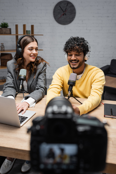 bearded indian man in headphones and yellow jumper talking in microphone near notebook and smiling colleague using laptop in front of digital camera recording podcast on blurred foreground - Foto, afbeelding
