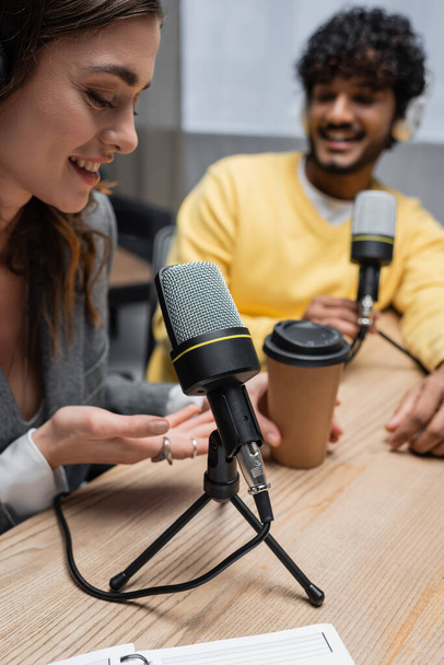 felice podcaster indicando drink da asporto vicino a microfoni professionali e collega indiano in maglione giallo sorridente su sfondo sfocato in studio radiofonico - Foto, immagini