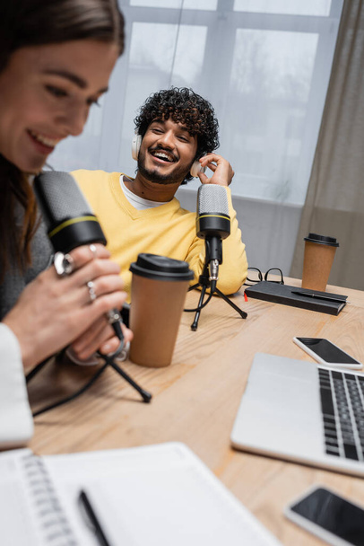 excited indian man in headphones and yellow jumper laughing near blurred colleague talking in microphone close to paper cups, notebooks, laptop and smartphone with blank screen in broadcasting studio - Photo, Image