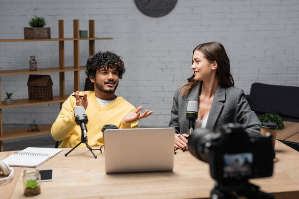 positive indian man in yellow jumper gesturing near microphones, laptop, notebook and smartphone while talking to smiling colleague sitting in blazer in front of blurred digital camera in studio - 写真・画像
