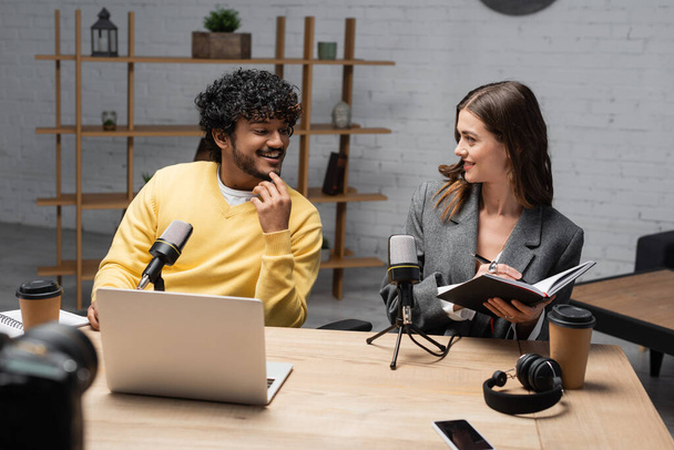 smiling brunette woman in grey blazer writing in notebook close to young and cheerful indian man in yellow jumper near microphones, laptop, coffee to go, headphones and smartphone in studio - Φωτογραφία, εικόνα