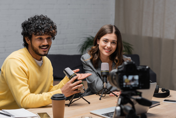 joyful and curly indian man in yellow jumper talking in microphone and pointing at charming colleague near laptop, coffee to go and smartphones in front of blurred digital camera in broadcasting studio - Photo, Image