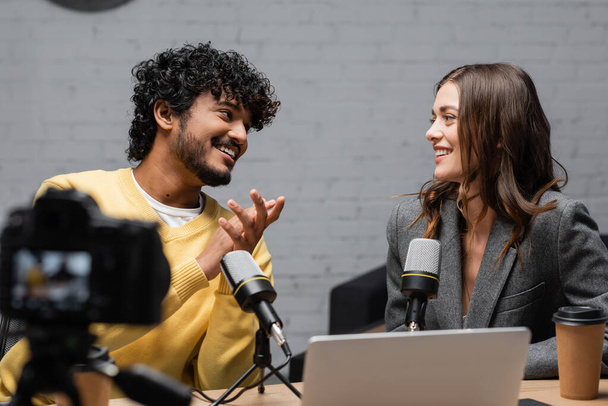 curly and bearded indian man in yellow jumper smiling at charming colleague sitting in grey blazer near microphones, laptop and coffee to go in front of blurred digital camera in studio - Photo, Image