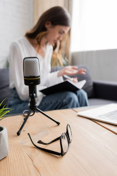 selective focus of table with laptop and eyeglasses and professional microphone near podcaster sitting on couch and working with notebook on blurred background - Photo, Image