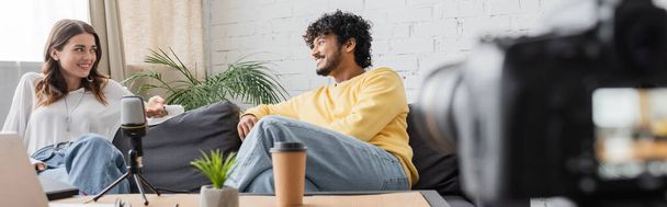 charming brunette woman and curly indian man in yellow jumper talking on couch near microphone and blurred paper cup with flowerpot while recording podcast on blurred digital camera, banner - Fotó, kép