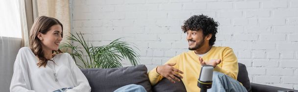 homem indiano encaracolado em amarelo jumper gesto enquanto sentado no sofá no estúdio de rádio e falando com mulher morena sorridente em blusa branca perto de planta verde contra parede de tijolo, banner - Foto, Imagem