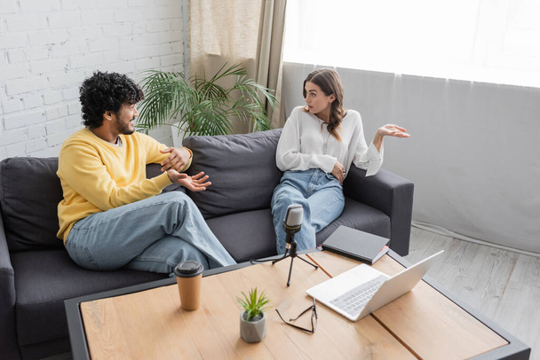 high angle view of multiethnic couple in casual clothes gesturing and talking on sofa near table with laptop, microphone, coffee to go, eyeglasses and flowerpot in radio studio - Photo, Image
