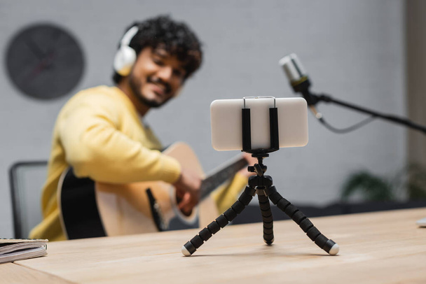 focus on smartphone on tripod near blurred indian musician in yellow jumper and headphones playing acoustic guitar close to professional microphone in broadcasting studio - Fotografie, Obrázek
