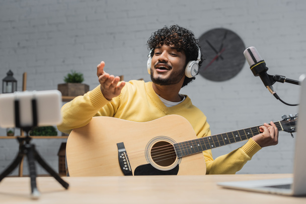 happy and curly indian podcaster in headphones and yellow jumper holding acoustic guitar and pointing with hand near blurred laptop and smartphone on tripod in record studio - Photo, Image