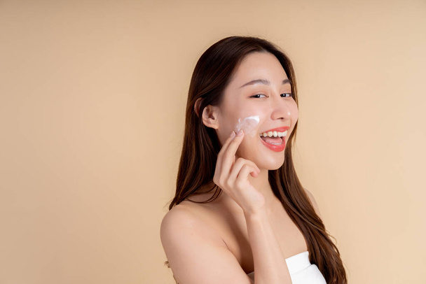 Nahaufnahme Porträt eines Gesichts des jungen asiatischen Mädchens mit gesunder Gesichtshaut, das sich vor Studiohintergrund mit weißer Gesichtscreme reibt. Hautpflege und dermatologisches Konzept. - Foto, Bild