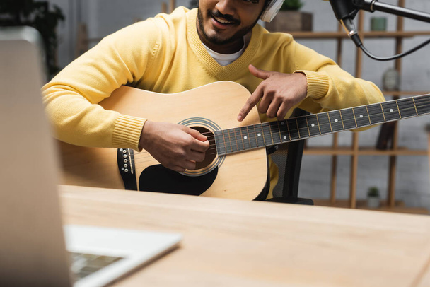 bijgesneden beeld van lachende indiaanse man in gele trui wijzend met de vinger naar akoestische gitaar bijna wazig laptop op houten tafel in omroepstudio - Foto, afbeelding