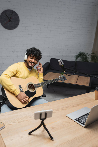Sonriente joven locutor indio en auriculares inalámbricos con guitarra acústica cerca de micrófono, teléfono inteligente y portátil durante la transmisión en el estudio  - Foto, imagen