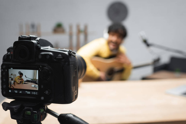 екран цифрової камери на тринозі, що стоїть біля розмитого молодого індійського підкастера, що тримає акустичну гітару біля мікрофона та ноутбука на столі в студії подкастів
  - Фото, зображення