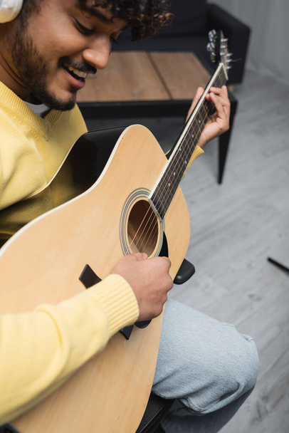 Blurred and smiling young indian musician in casual clothes and wireless headphones singing and playing acoustic guitar during performance in studio  - 写真・画像