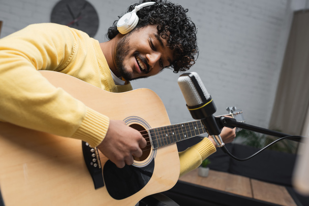 Positive und junge indische Podcaster mit Kopfhörern spielen Akustikgitarre in der Nähe von Mikrofon auf Ständer während Stream im Podcast-Studio  - Foto, Bild