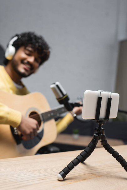 Smartphone em tripé na mesa perto de podcaster indiana borrada em fones de ouvido tocando guitarra acústica perto de microfone durante fluxo em estúdio de podcast, smartphone em tripé  - Foto, Imagem