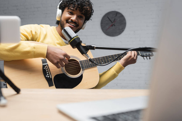 Emissora indiana sorridente em fones de ouvido sem fio tocando guitarra acústica durante a performance perto do microfone e laptop borrado na mesa em estúdio  - Foto, Imagem