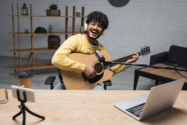 Carefree giovane podcaster indiano in cuffie wireless suonare la chitarra acustica vicino a dispositivi e caffè per andare in tazza di carta e quaderno sul tavolo in studio podcast  - Foto, immagini