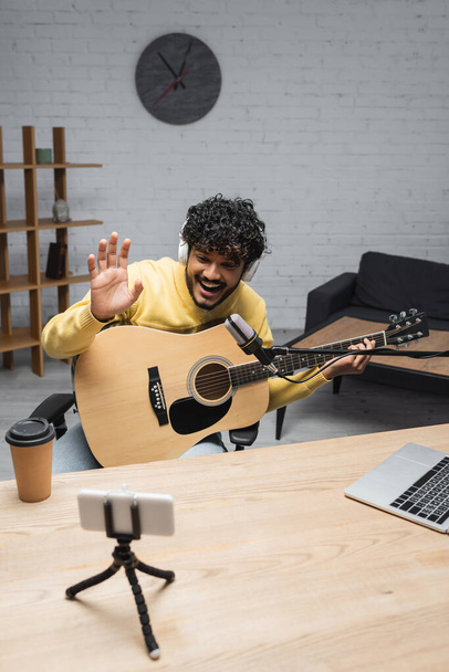 Junge und positive indische Podcaster in drahtlosen Kopfhörern winken während eines Videogesprächs mit dem Smartphone und halten eine Akustikgitarre in der Nähe von Coffee to go und einen Laptop auf dem Tisch im Podcast-Studio  - Foto, Bild