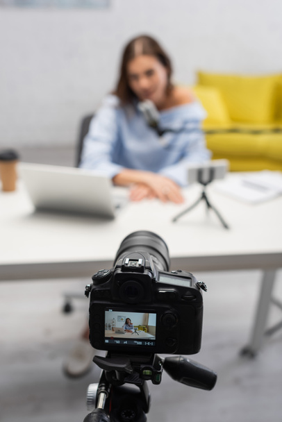 screen of digital camera on tripod standing near blurred brunette blogger talking at microphone near laptop on table during stream in studio  - 写真・画像